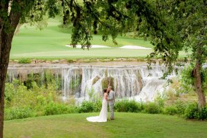 bride and groom standing in front of a waterfall at The Hills Country Club