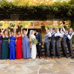 Wedding party under pergola, bridesmaids in multi-colored dresses, men in suits, all cheering